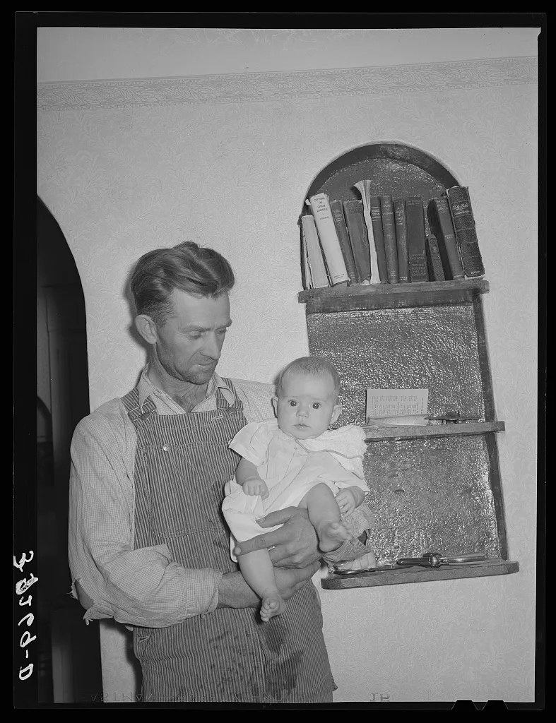 Mormon farmer with his baby in front of a bookshelf in his home.…, Utah, 1940