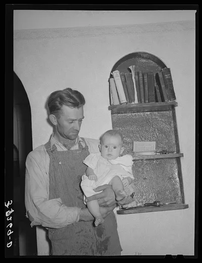 Mormon farmer with his baby in front of a bookshelf in his home.…, Utah, 1940